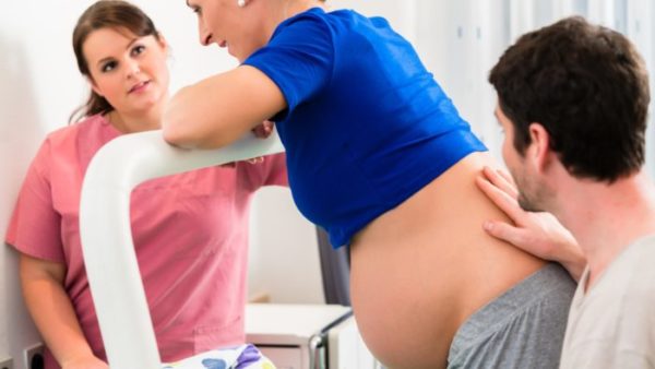 ngo-illustration-Artboard 5 Tehotná žena kľačí na posteli a operia sa o podpornú tyč. Partner ju hladí po krížoch. Pôrodná asistentka je pri nej a pozerá sa jej príamo do tváre. Právo na sprevádzajúcu osobu je stanovené zákonom.