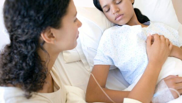 Staršie dievča, leží v nemocničnej posteli, v jednej ruke má infúziu, druhú ruku má položenú na hrudi. Mama sedí vedľa nej a má tiež položenú ruku na jej ruke na hrudníku.